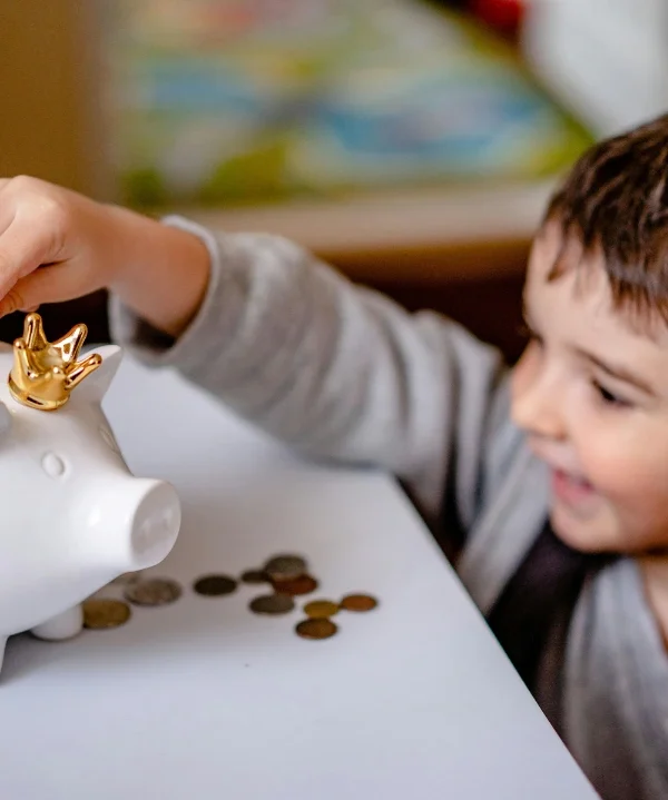 Young boy happily putting coins into a piggy bank, illustrating Dr. Siggie's spend save share approach to teaching child allowance spending