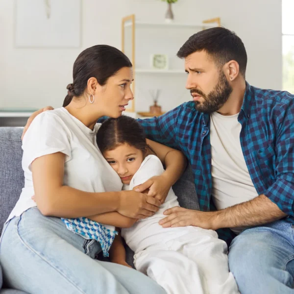 Two parents sitting on a couch with their young daughter between them, arguing over parenting decisions while the child looks distressed — illustrating the good cop bad cop parenting